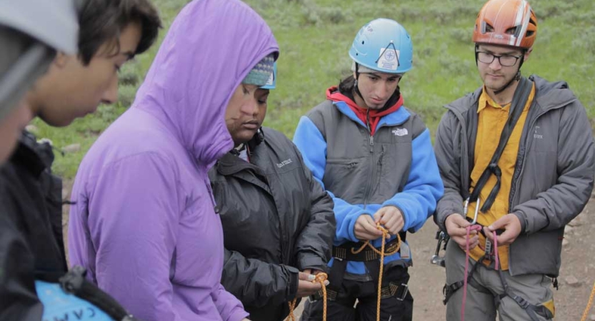 A group of people wearing safety gear appear to practice tying knots. 